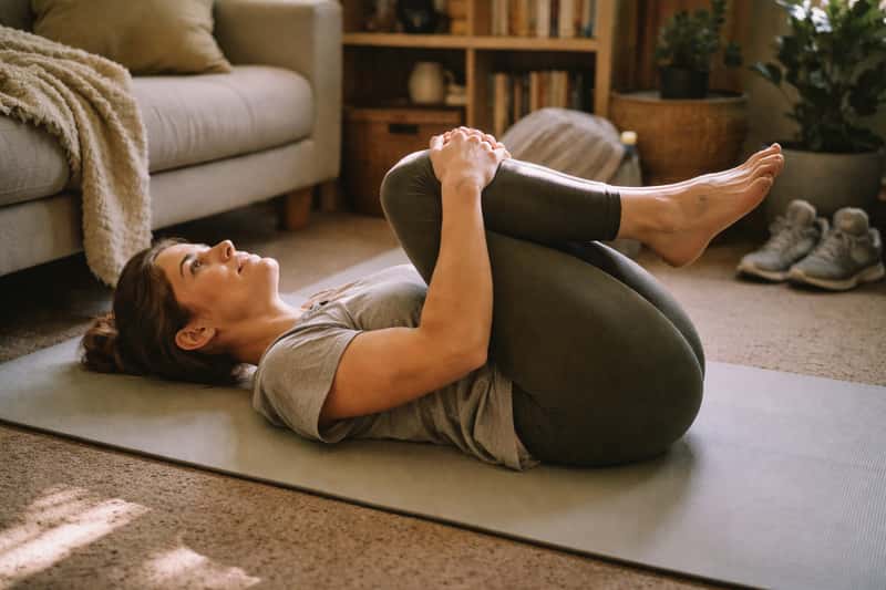 Person lying on back with one knee pulled toward chest, other leg extended, demonstrating stretching position on wooden floor