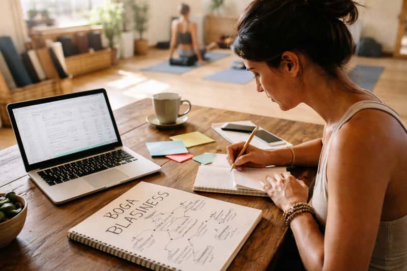 Yoga instructor reviewing business documents and laptop at Yoga instructor reviewing business documents and laptop at studio desk with water bottle and notebook
